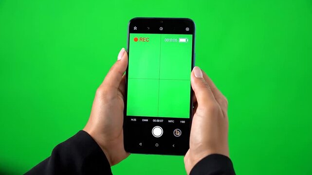 A person holding a smartphone filming against a green screen. The phone displays a video recording interface