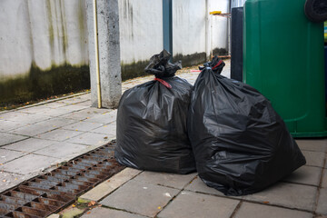 Two large, black trash bags sit tied up beside a green bin and a rusted drain grate.