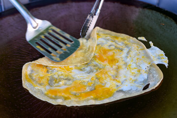 An egg crepe is cooked on a hot griddle, being folded with a spatula and tongs.