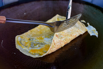 An egg crepe is cooked on a hot griddle, being folded with a spatula and tongs.