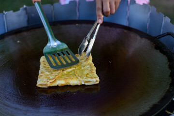 An egg crepe is cooked on a hot griddle, being folded with a spatula and tongs.