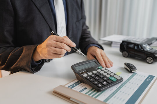 A businessman in a suit is calculating car loan options using a calculator and comparison chart, with model cars and car keys on the desk, symbolizing finance, leasing, and investment planning.