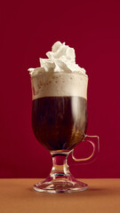 Irish coffee with whipped cream topping served in clear glass on red background. Concept of dessert beverage, cafe advertising, festive branding, and luxurious lifestyle indulgence.
