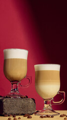 Two layered latte macchiato glasses on red background with roasted coffee beans. Concept of beverage composition, cafe branding, product display, and luxury culinary design.