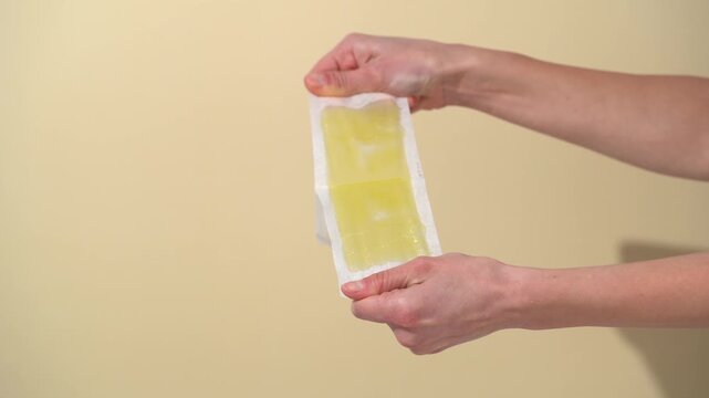 Female hands uncover two pieces of yellow wax strips for depilation on a beige background. Concept of waxing strips relies on natural resins and oils for effective adhesion and skin care.