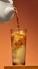 Coffee poured from white pot into tall glass creating swirl pattern over brown background. Concept of beverage design, culinary lifestyle, cafe branding, and modern advertising scene.