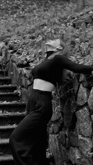 A woman in a black crop top and wide-leg pants stands by a stone wall on the steps in a park. She has her head thrown back and is relaxedly breathing in the fresh air, enjoying a moment of peace.