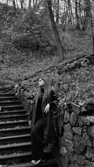 A woman in a black crop top and wide-leg pants stands by a stone wall on the steps in a park. She has her head thrown back and is relaxedly breathing in the fresh air, enjoying a moment of peace.