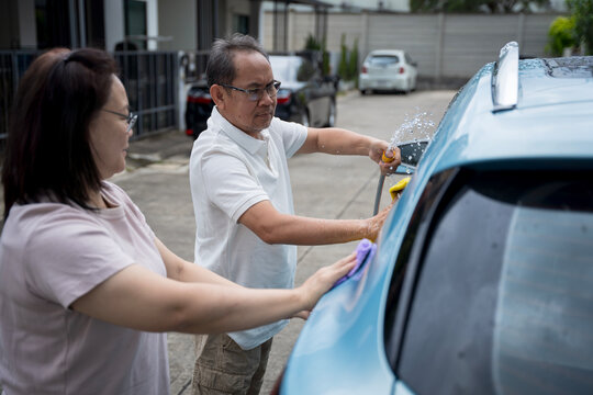 Middle aged Asian couple enjoys quality time together washing their blue car on a sunny day