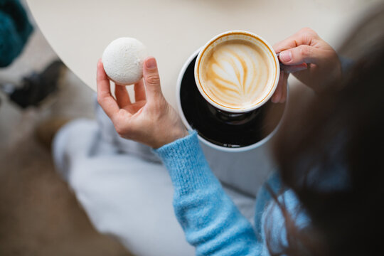Overhead view of a woman sitting at a cafe drinking a cappuccino and eating a vanilla and coconut macaroon