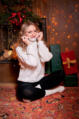 A beautiful girl was sitting on the floor, surrounded by gifts that were presented to her in honor of the holiday. She is wearing a white sweater and dark trousers. The trend of Christmas celebrations