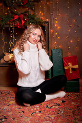 A beautiful girl was sitting on the floor, surrounded by gifts that were presented to her in honor of the holiday. She is wearing a white sweater and dark trousers. The trend of Christmas celebrations