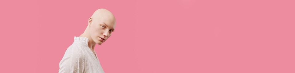 Website banner of young adult Caucasian woman with alopecia looking into camera, standing against pink background, wearing light colored top, neutral facial expression