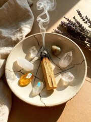 A ceramic plate holds a piece of wood with smoke rising, surrounded by various crystals and lavender sprigs. The scene conveys a calming, spiritual atmosphere.