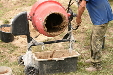A man in a blue T-shirt and green pants turns the handle of a red cement mixer to pour freshly mixed clay into a container on green grass. Outdoor construction or DIY work scene.