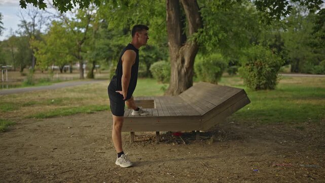 A fit man performs hamstring stretches on a wooden bench in a lush green park. This outdoor exercise promotes health, flexibility, and a healthy lifestyle.