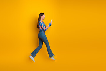 Young woman walking and looking at her phone against a vibrant yellow background capturing casual...