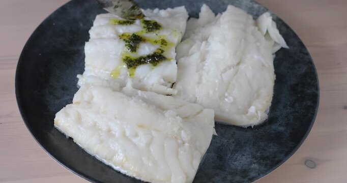 Close-up of three boiled cod fillets on a gray plate as a spoon drizzles olive oil mixed with parsley over the fish. Healthy eating, Mediterranean, traditional food, and simple home cooking. No audio.