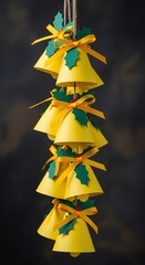 Vertical arrangement of paper craft yellow bells decorated with holly leaves and orange ribbons, artistic festive ornament hanging on dark blurred background