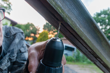 An older man drills into a weathered wooden beam outdoors, working on a home improvement project...