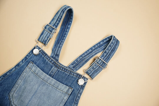 A pair of denim overalls rests on a soft beige surface, highlighting the straps and pocket design. The fabric texture and stitching are clearly visible, emphasizing the casual style.