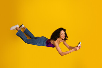 Young woman in chic casual fashion floats holding smartphone against bright yellow background creating a fun lifestyle moment