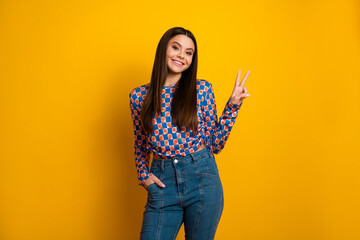 Vibrant young woman in checkered top posing with peace sign against a bright yellow background...