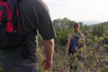 A couple with backpacks walks downhill along a natural trail surrounded by bushes and trees,...