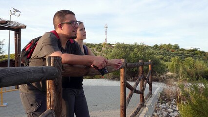 Couple leaning on a wooden fence, resting after hiking through a green landscape of olive trees. Concept of relaxation, connection, and outdoor lifestyle.