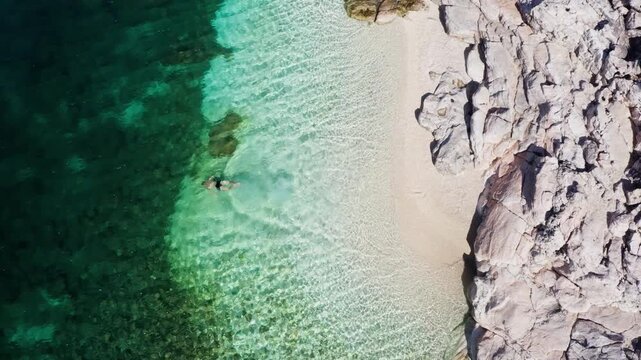 Top view 4K drone video of man dressed black swimming trunks swimming relaxing on white pebble lonely beach water waves. Exotic tropical summer holidays concept. Cephalonia island, Greece.