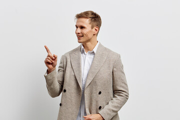Confident young man in stylish beige blazer and white shirt pointing with finger while smiling, standing against plain white background. Studio portrait, fashion, success, communication, professional