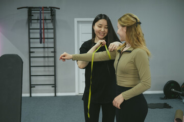 Two sporty women smiling and measuring their body parameters with a yellow measuring tape in a cozy sport room, celebrating their fitness progress after an intense workout session.