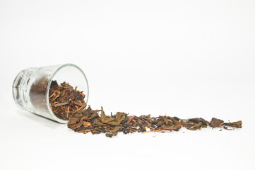 Small glass cup tipped over with dried loose tea leaves spilled out onto a pure white background. Represents tea, herbal beverage, health, natural ingredient, isolated, and minimalist food concept.