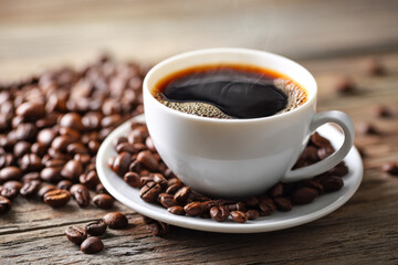 Obraz premium Hot steaming black coffee in a white ceramic cup surrounded by roasted coffee beans on a rustic wooden table with natural warm lighting and cozy ambiance