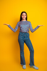 Bright young woman with patterned top and jeans stands against a yellow background smiling with...