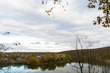 Autumn trees with yellow and red leaves on a hill above calm river reflecting forest and cloudy sky. Rural landscape photography. Autumn season and nature concept.