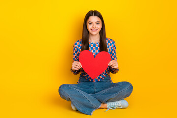 Cheerful young girl sits cross legged holding a red heart against yellow backdrop expressing love...
