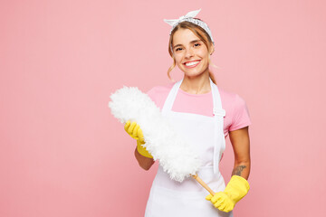Young smiling cheerful woman wears white apron hold use feather duster look camera while doing housework tidy up isolated on plain pastel light pink background studio portrait. Housekeeping concept. © ViDi Studio