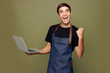 Young IT man barista bartender barman in jeans apron black t-shirt work in coffee shop hold use laptop pc computer do winner gesture isolated on plain green background Small business startup concept