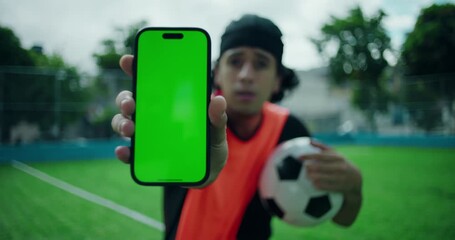 Hispanic man holding smartphone with green screen toward camera while carrying soccer ball, presenting mobile app or sports concept with focus on technology - Powered by Adobe