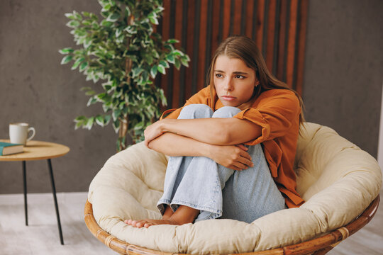 Fototapeta Full body young sad depressed frustrated woman wear orange casual clothes hold knees sits in chair stay at home hotel flat rest relax spend free spare time in living room indoor. People lounge concept