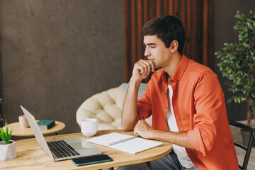 Young thoughtful IT man wear casual clothes work or study on laptop pc computer sit alone at table in coffee shop cafe relax rest in restaurant during free time indoor Freelance mobile office concept