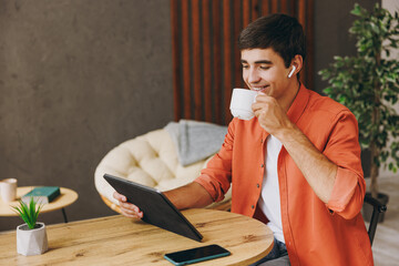 Young IT man he wear orange casual clothes digital tablet pc computer drink tea sit at table in coffee shop cafe relax rest in restaurant in free time indoor. Freelance mobile office business concept