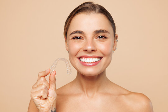 Half naked topless smiling fun young woman 20s with nude make up hold invisible transparent aligners braces isolated on plain beige background studio. Skin care healthcare cosmetic procedures concept.