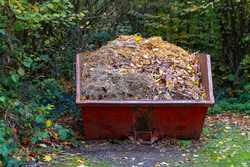 Bright red container filled with garden waste surrounded by autumn leaves in a serene forest setting