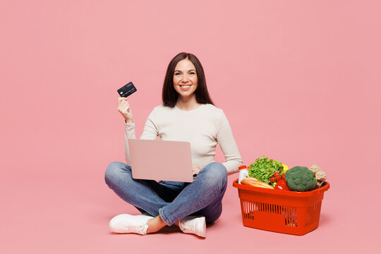 Full body young IT woman wear white casual clothes sit near basket bag food products hold credit bank card use work on laptop pc computer isolated on plain pink background. Delivery service from shop.