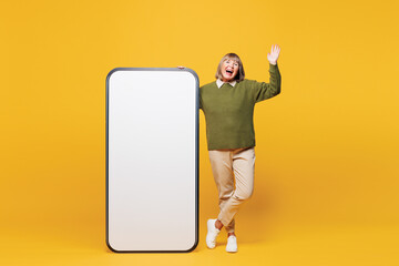 Full body elderly smiling happy woman wear green sweater casual clothes big huge blank screen area mobile cell phone smartphone spread hand isolated on plain yellow orange background studio portrait.