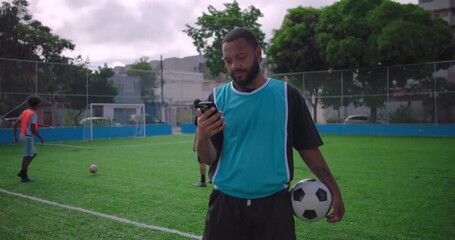 Hispanic man standing on soccer field checking smartphone while holding ball, showing focus and casual interaction with technology during sports practice outdoors - Powered by Adobe