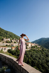 Naklejka premium Beautiful elegant woman in a light rose dress walking gracefully in front of the spectacular view of Cargèse village Corsica, France. Scenic Mediterranean landscape with charming coastal architecture