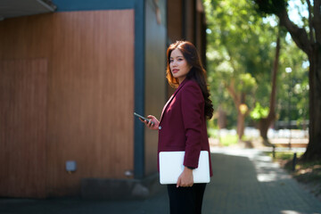 Asian businesswoman walking outdoors holding phone and laptop